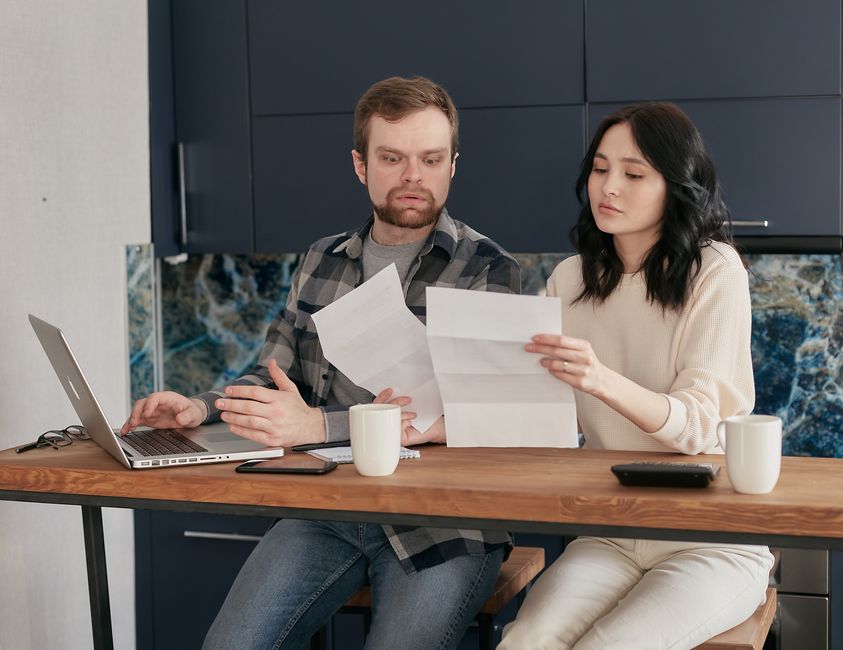 A couple sitting at their kitchen table, looking at a laptop screen together, discussing their household budget with an open finance application visible, emphasizing financial planning.