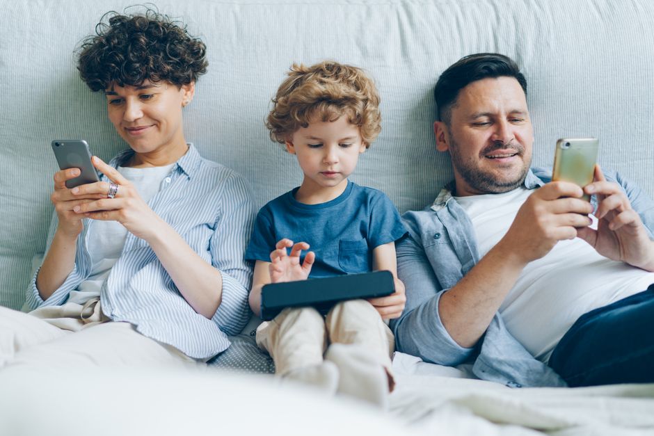 A family, including parents and children, gathered around a table with multiple devices like laptops, phones, and tablets, with a glowing VPN shield icon connecting them all.