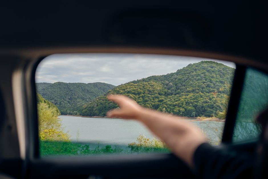Travelers enjoying a break during a road trip, with hands reaching for healthy travel snacks like fruit and sandwiches from a cooler in a car with a beautiful landscape visible outside.
