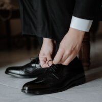 A close-up of a man's hands tying the laces of a polished brown leather Oxford shoe.