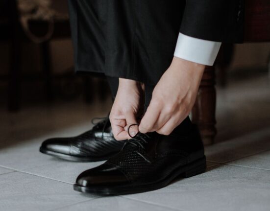 A close-up of a man's hands tying the laces of a polished brown leather Oxford shoe.