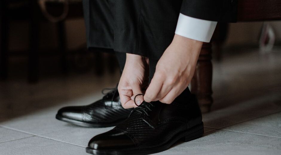 A close-up of a man's hands tying the laces of a polished brown leather Oxford shoe.