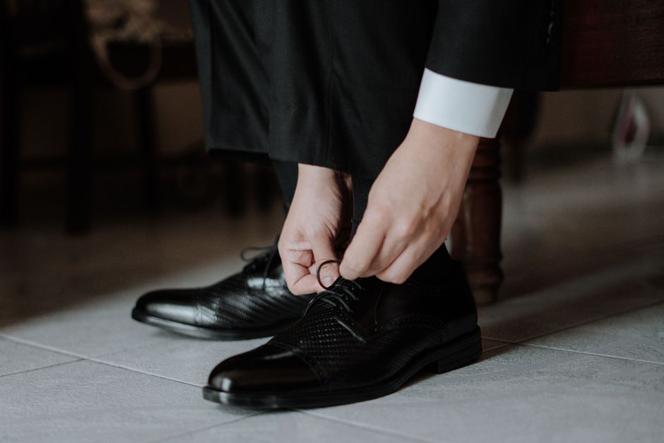 A close-up of a man's hands tying the laces of a polished brown leather Oxford shoe.