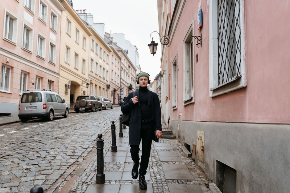 A stylish man in smart casual attire walking confidently down a historic, cobbled European city street, carrying a small messenger bag.