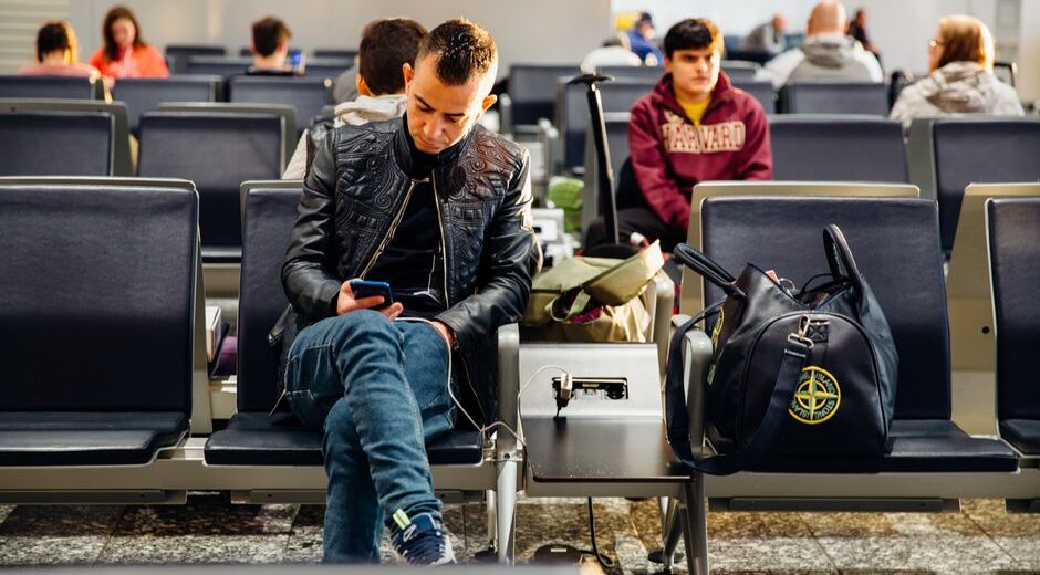 A traveler charging their smartphone with a compact portable power bank at an airport gate, with other luggage in the background.