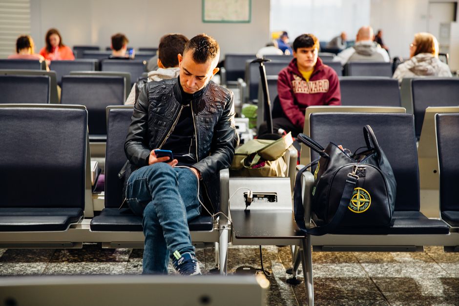 A traveler charging their smartphone with a compact portable power bank at an airport gate, with other luggage in the background.