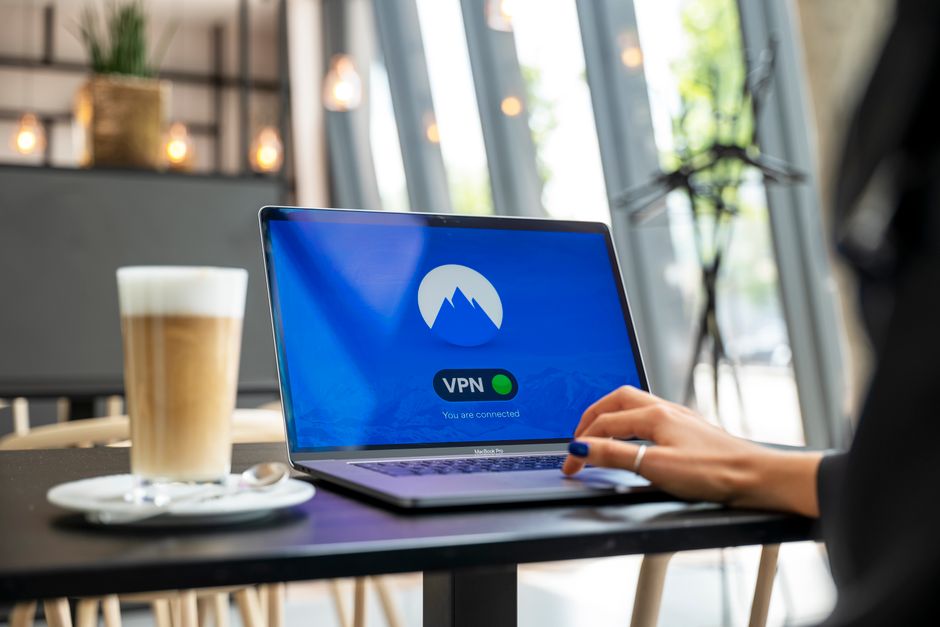 A person using a laptop at a cafe table, with a glowing shield icon overlaid on the screen, indicating a secure connection on public Wi-Fi.