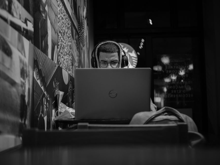 A person wearing large noise-cancelling headphones concentrating at a computer in a modern home office, with a clear webcam visible.