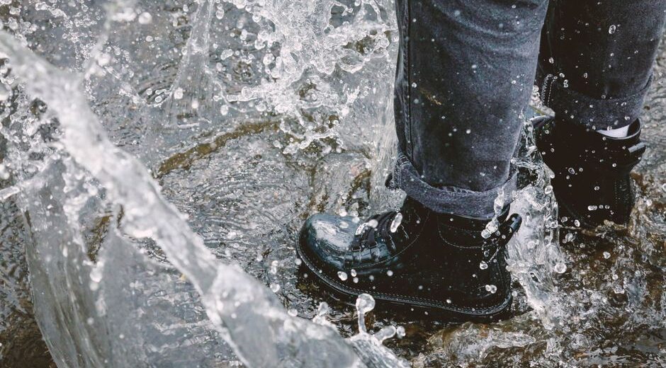 A person wearing dark waterproof walking shoes, walking confidently on a wet city street in the rain, with puddles visible.