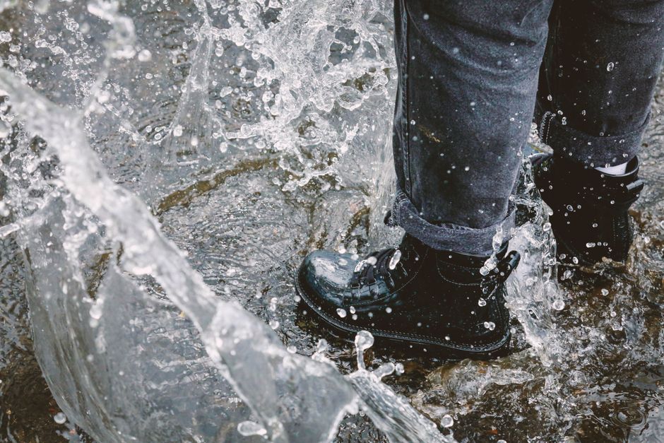 A person wearing dark waterproof walking shoes, walking confidently on a wet city street in the rain, with puddles visible.