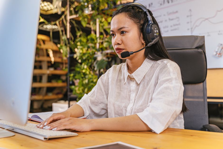 A focused individual wearing over-ear noise-cancelling headphones, sitting at a home office desk with a laptop and monitor setup.