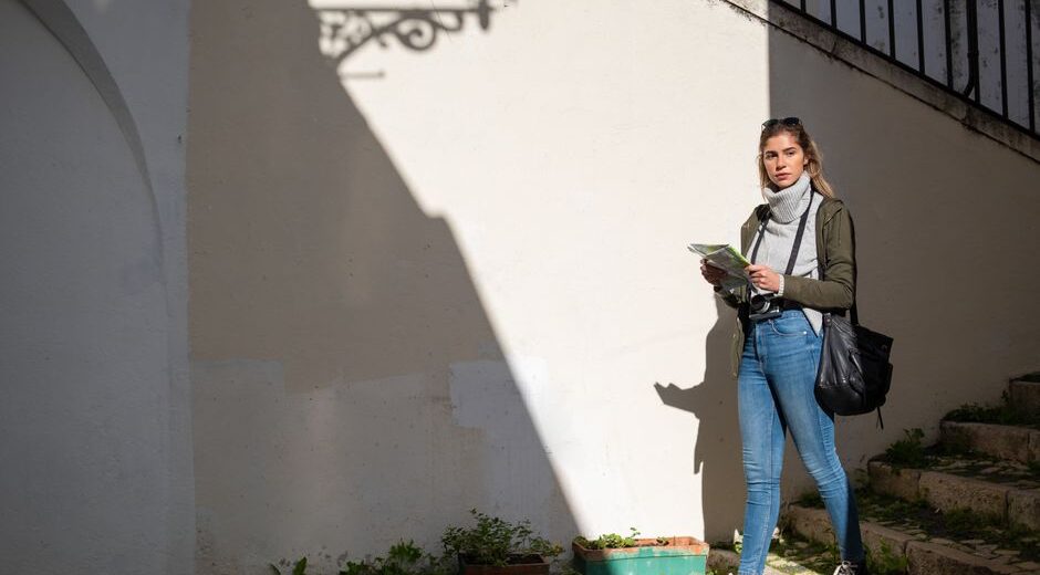 A solo female traveler in a smart casual outfit, holding a map and looking up at historic buildings in a busy European city street.