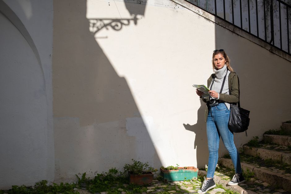 A solo female traveler in a smart casual outfit, holding a map and looking up at historic buildings in a busy European city street.