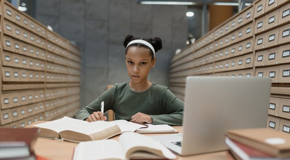 A female student thoughtfully comparing laptop models on a tablet, surrounded by notebooks and pens, embodying the tech buying process.