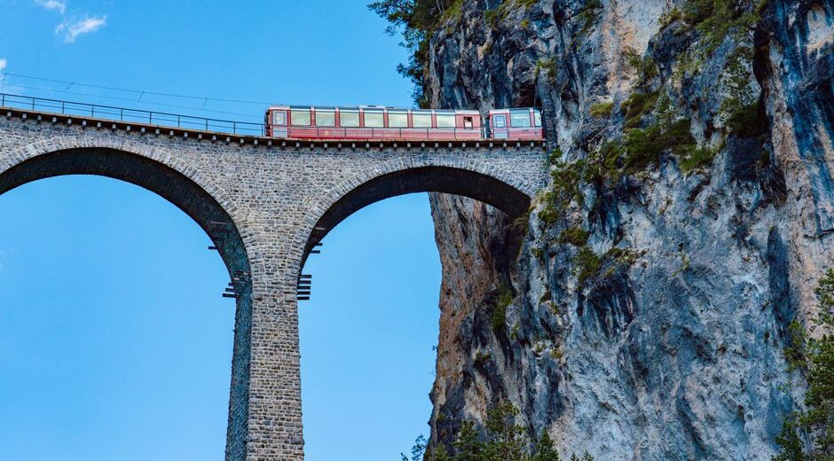 Red Bernina Express train crossing the Brusio spiral viaduct in the Swiss Alps, surrounded by green landscape.