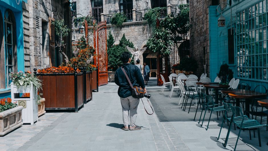 A traveler in a cafe securing a black anti-theft crossbody bag to the leg of a wooden chair using its integrated anchor strap, with a laptop and coffee on the table.