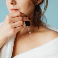 A woman gently patting a hydrating serum onto her face, which has water droplets visible, in a bright bathroom.