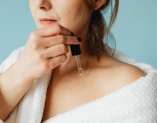 A woman gently patting a hydrating serum onto her face, which has water droplets visible, in a bright bathroom.