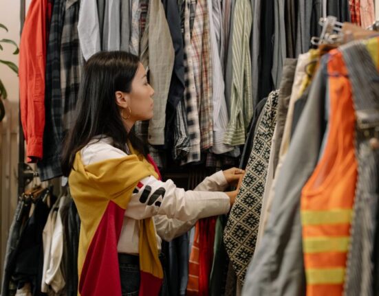 A woman thoughtfully examining a blouse on a hanger in a brightly lit, minimalist sustainable fashion store.