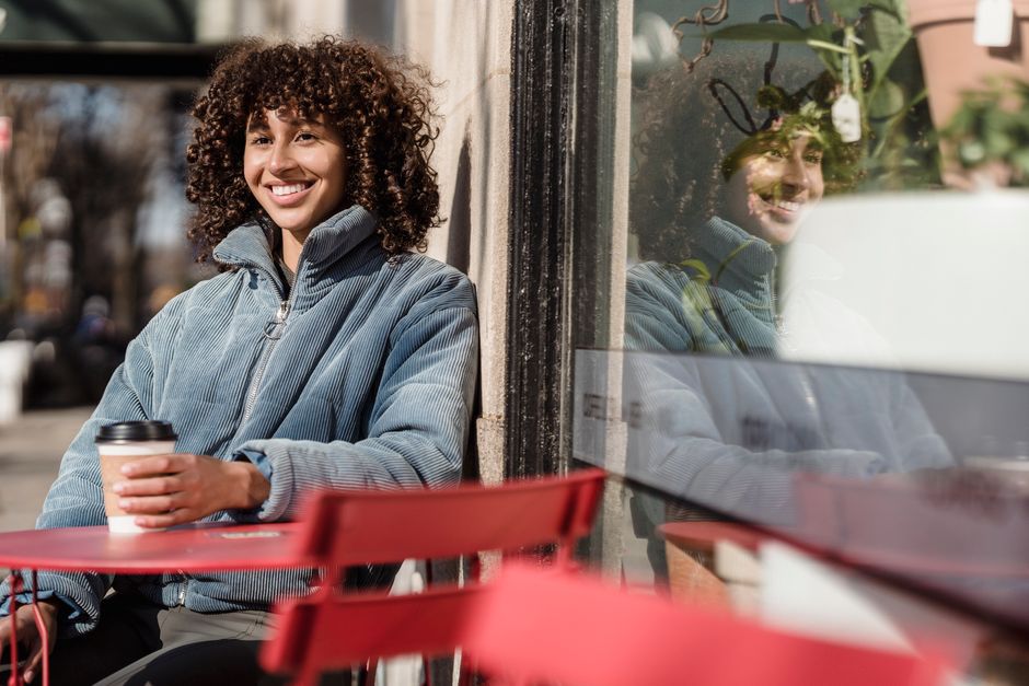 A woman smiling while sipping coffee at a small, authentic outdoor cafe, observing local life in a quiet square.