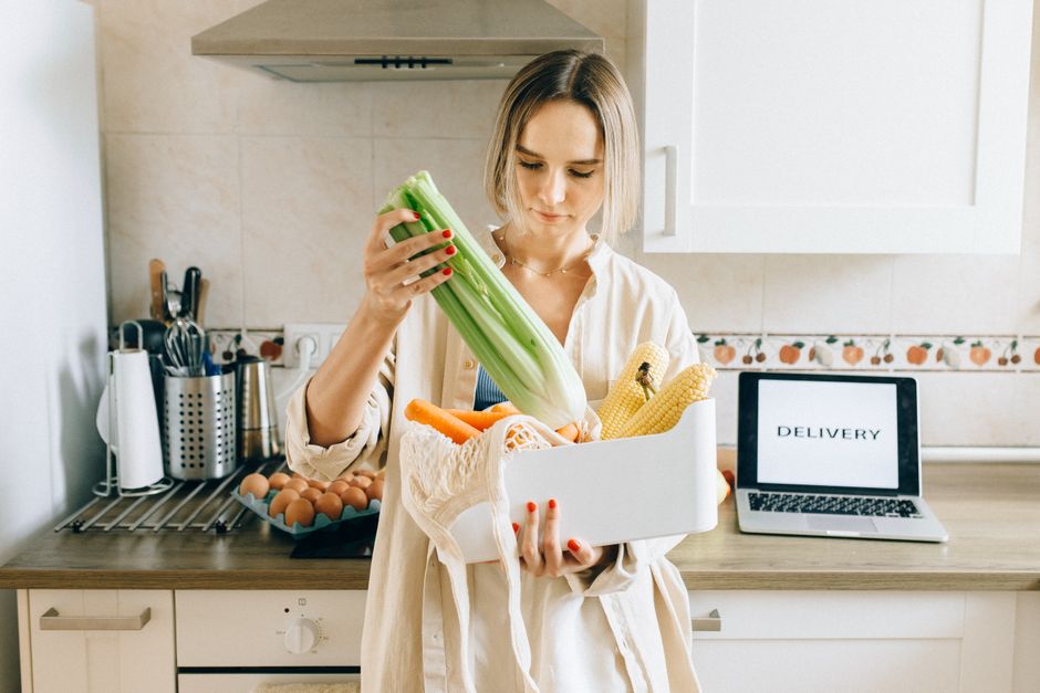 A woman smiling while sitting at her kitchen table, browsing for groceries on her laptop, with neatly stacked delivery boxes visible in the background, illustrating online shopping convenience.
