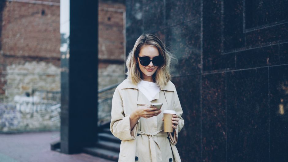 A woman wearing a classic trench coat, a light scarf, and holding a coffee cup, looking ready for a cool spring day, exemplifying smart layering for unpredictable spring weather.